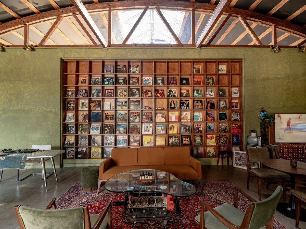 A mid-century modern lounge space with exposed wood beam ceiling, vinyl record display wall, tan leather sofa, vintage furniture, and an engine block 