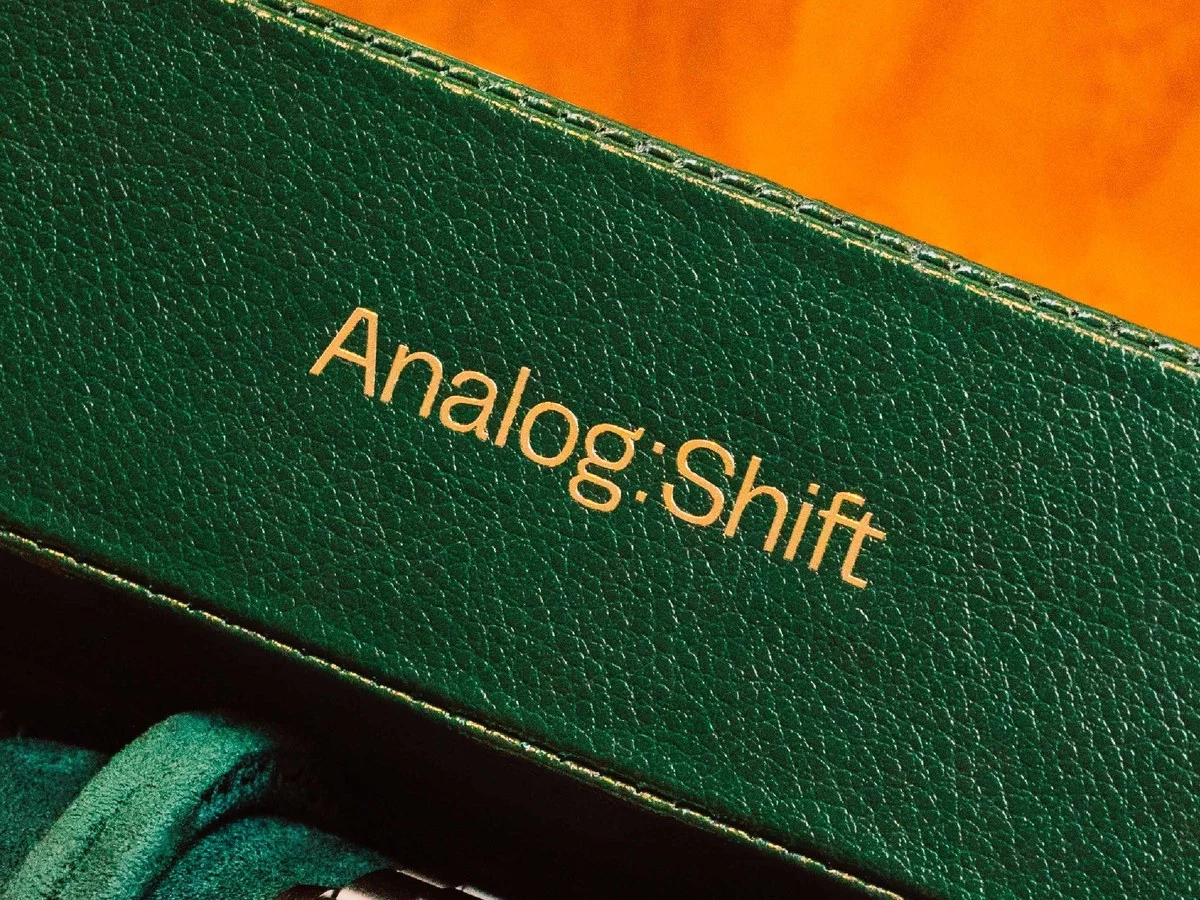 A green leather watch box with the gold text "Analog:Shift" embossed on its lid, partially revealing a Rolex watch inside.