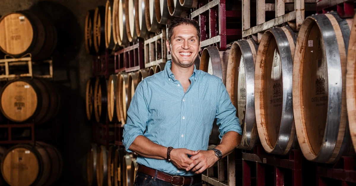 A man in a blue button-down shirt stands smiling in a wine barrel cellar