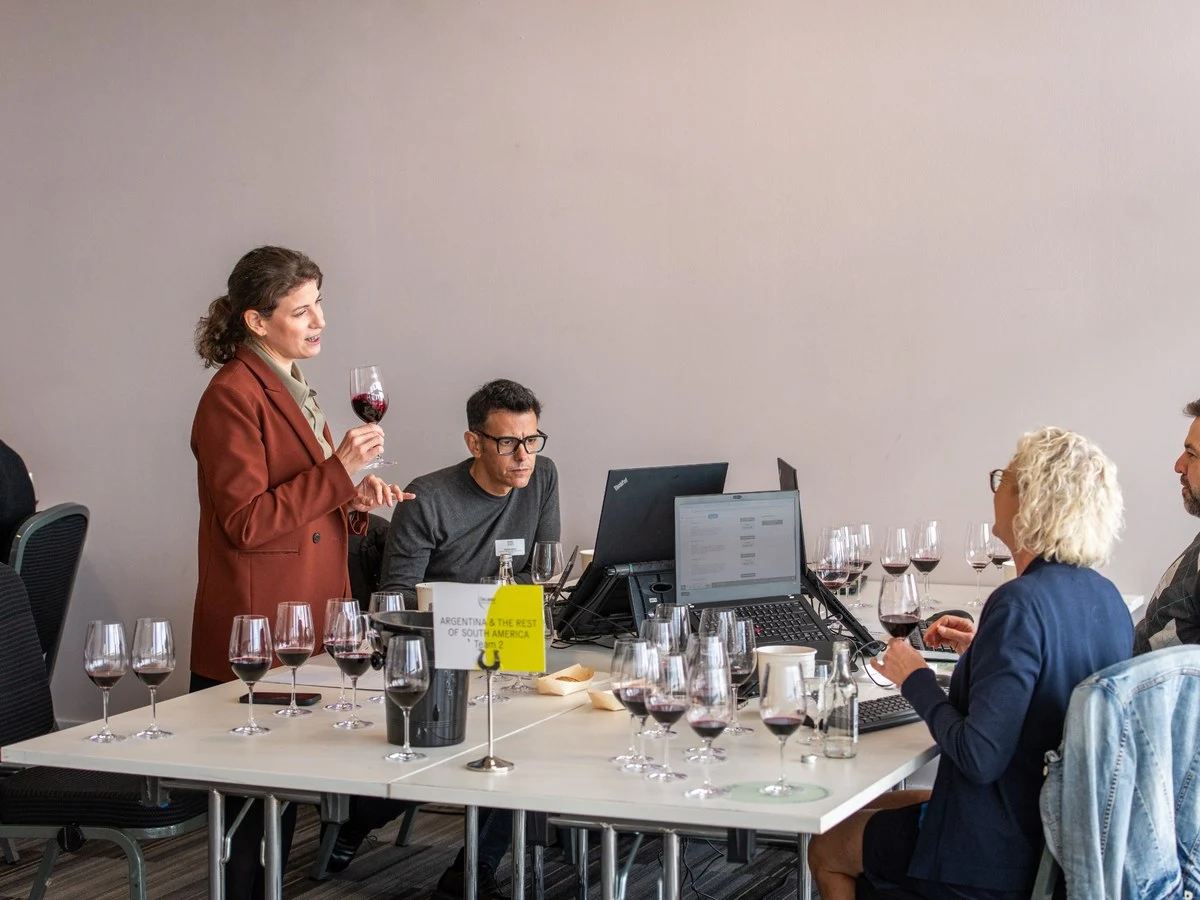 A wine tasting session with a woman in a rust-colored blazer standing and holding a wine glass, addressing three seated participants at a table with m