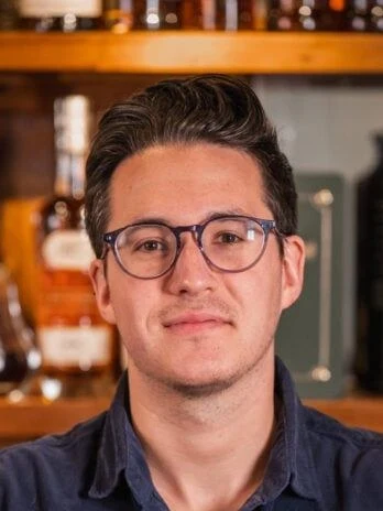 A man wearing glasses and a navy corduroy shirt stands behind a wooden bar with liquor bottles on shelves.