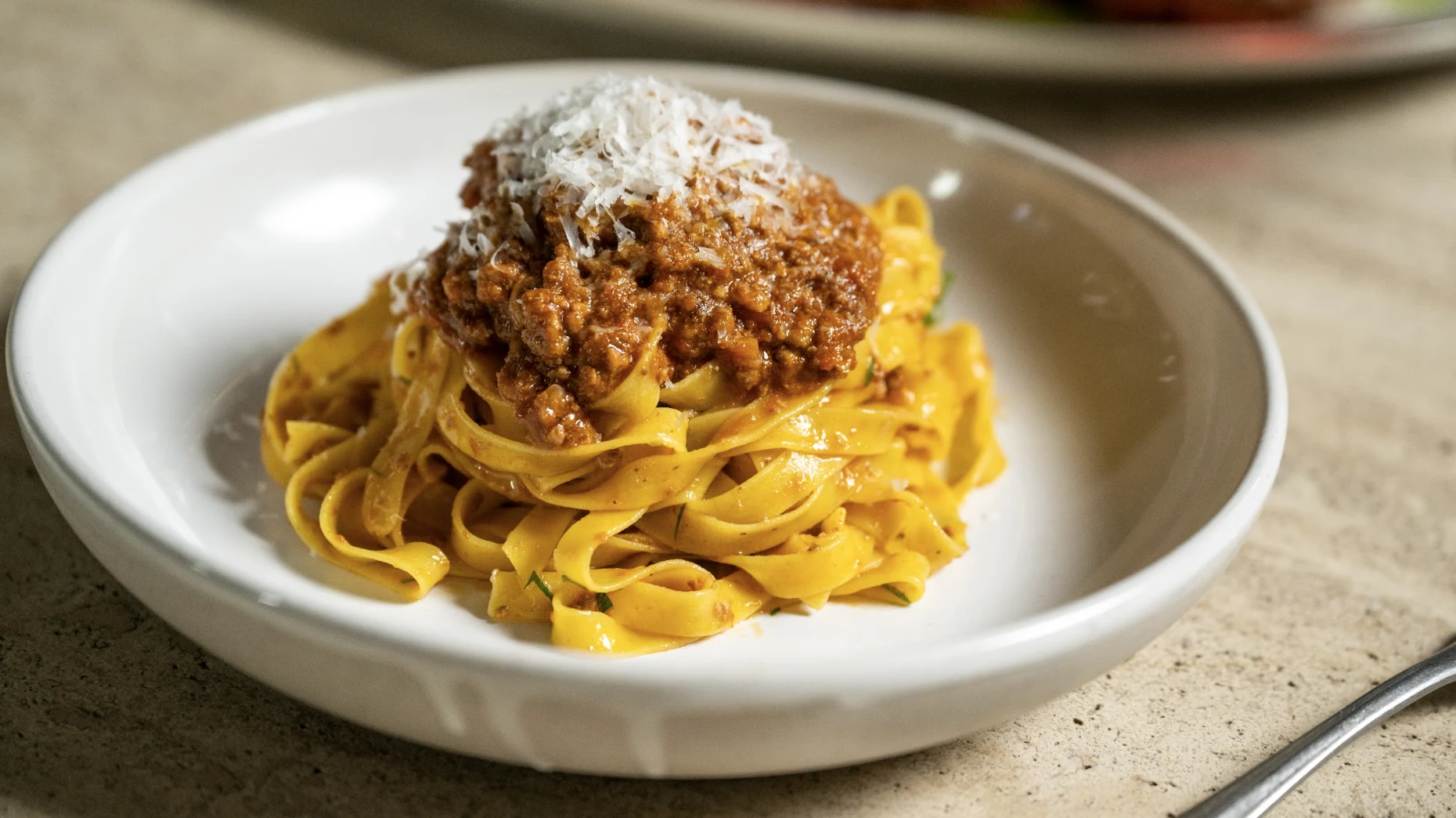 A white bowl containing yellow ribbon pasta topped with brown meat sauce and white shaved cheese on a concrete surface.