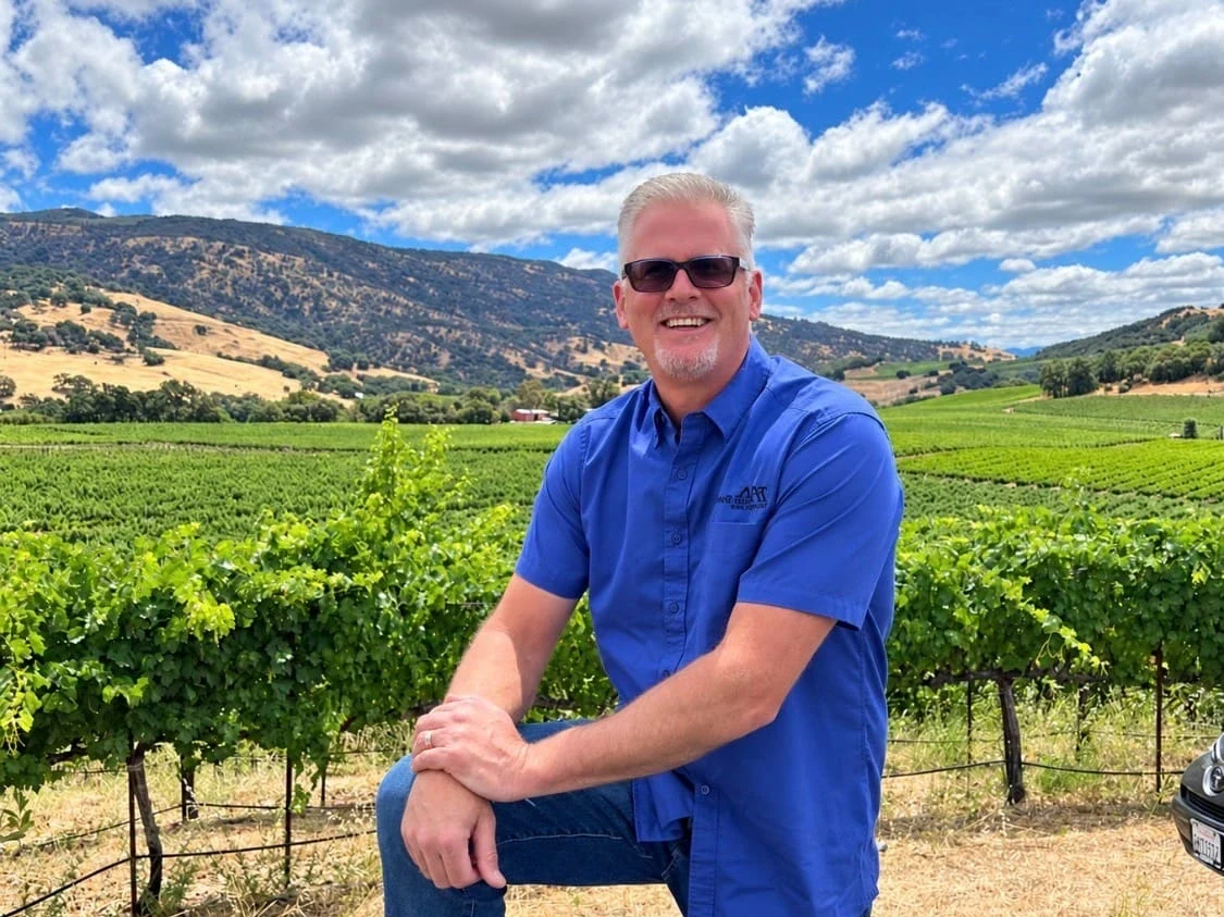 A man wearing a blue short-sleeved shirt and sunglasses stands in a vineyard, smiling at the camera with grapevines and hillside landscape behind him.