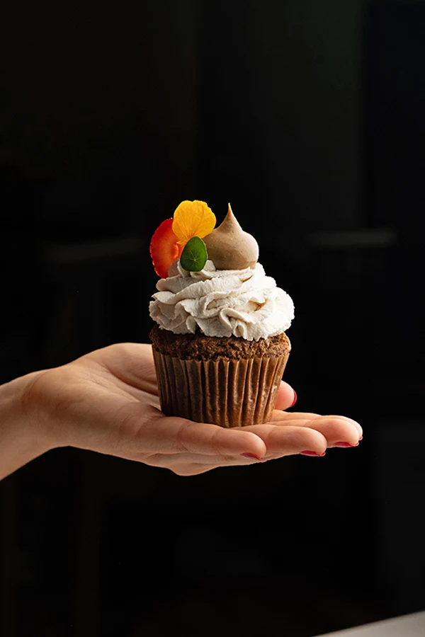 A hand holding an elegantly decorated cupcake with white frosting, topped with edible flowers and a meringue kiss; the source page explicitly attests 