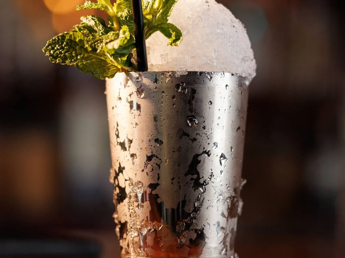 A close-up of a Mint Julep cocktail in a silver metal cup, overflowing with crushed ice and garnished with fresh green mint leaves, with a black.