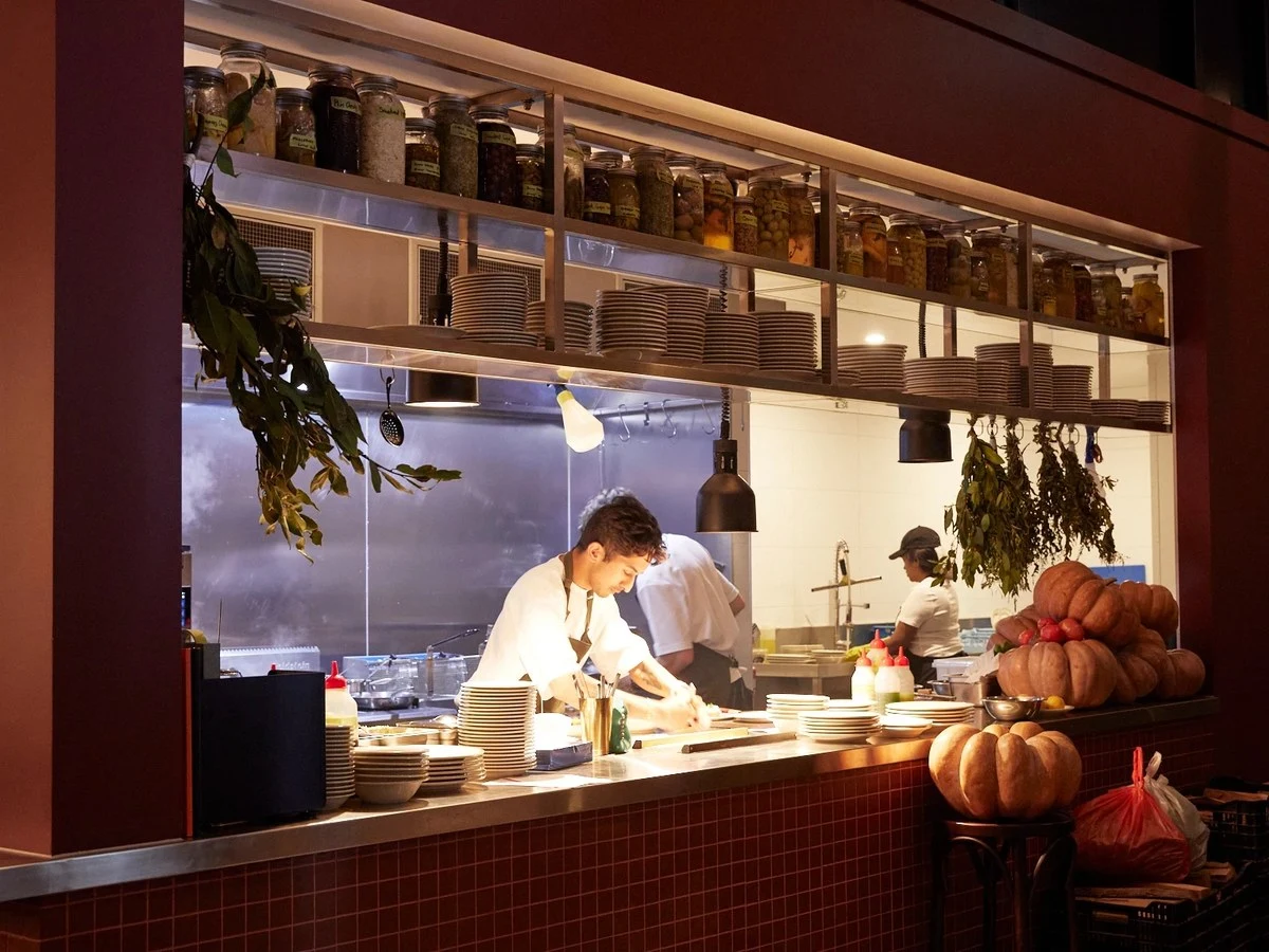 An open restaurant kitchen showing a chef in white uniform and brown apron leaning over a pass plating dishes, with stacked plates, hanging herbs, row