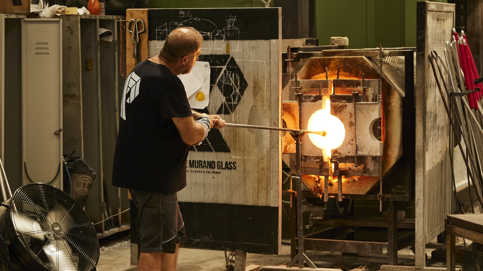 Glassblower working molten glass at a furnace inside a Murano glass studio, with glowing orange gather on blowpipe.