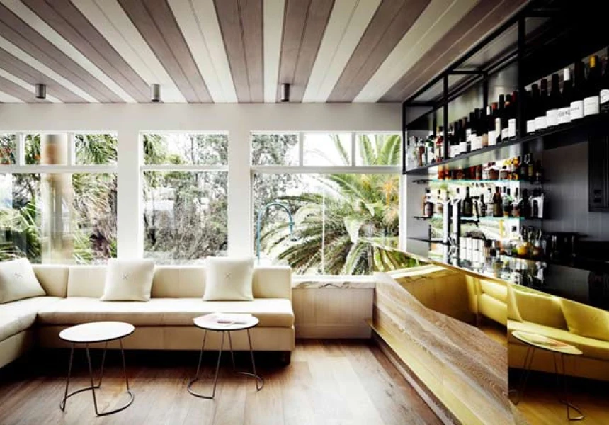 An upscale restaurant dining room interior with large windows overlooking tropical vegetation, cream-colored banquette seating, a yellow velvet sofa, 