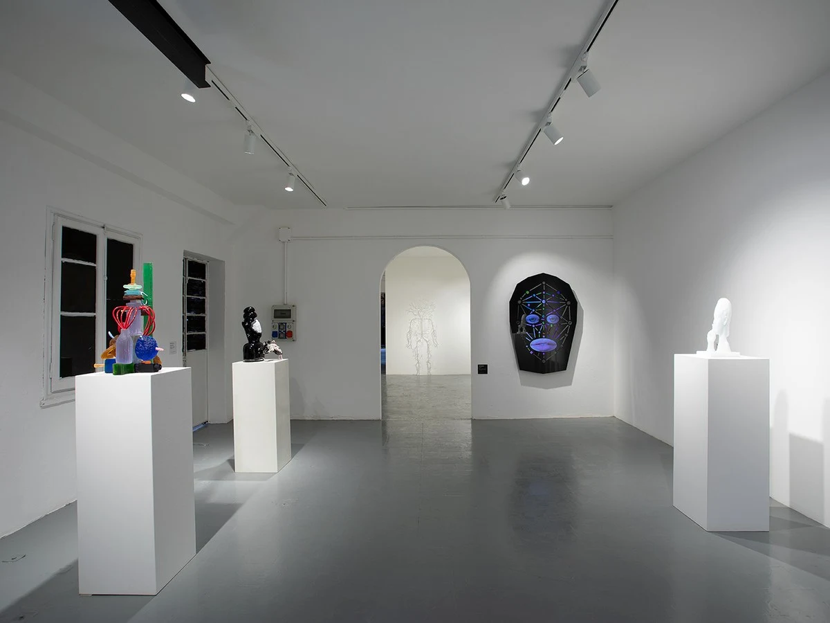 An interior view of a contemporary art gallery with white walls and grey floors, showcasing various glass sculptures on pedestals.