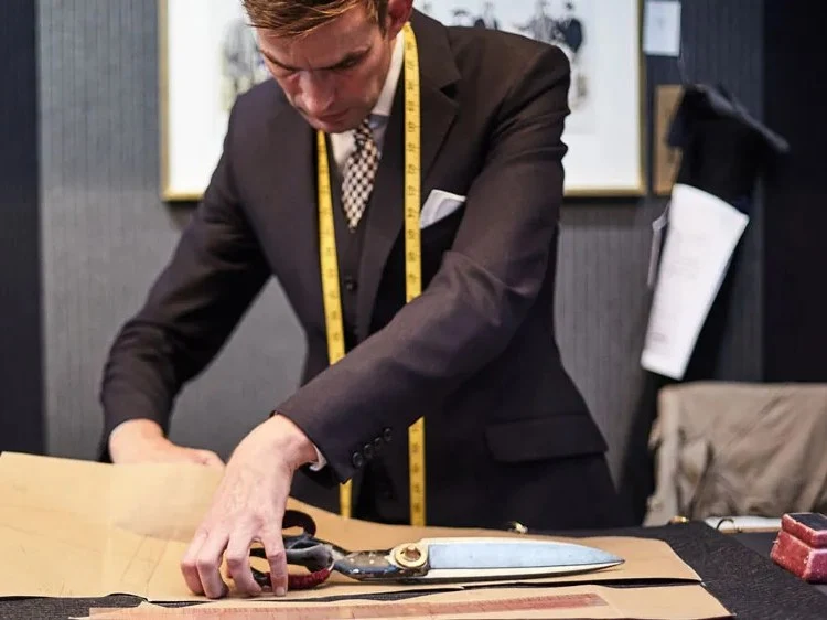 A tailor in a dark suit with a yellow measuring tape around his neck cuts a paper pattern on a workbench using large shears.