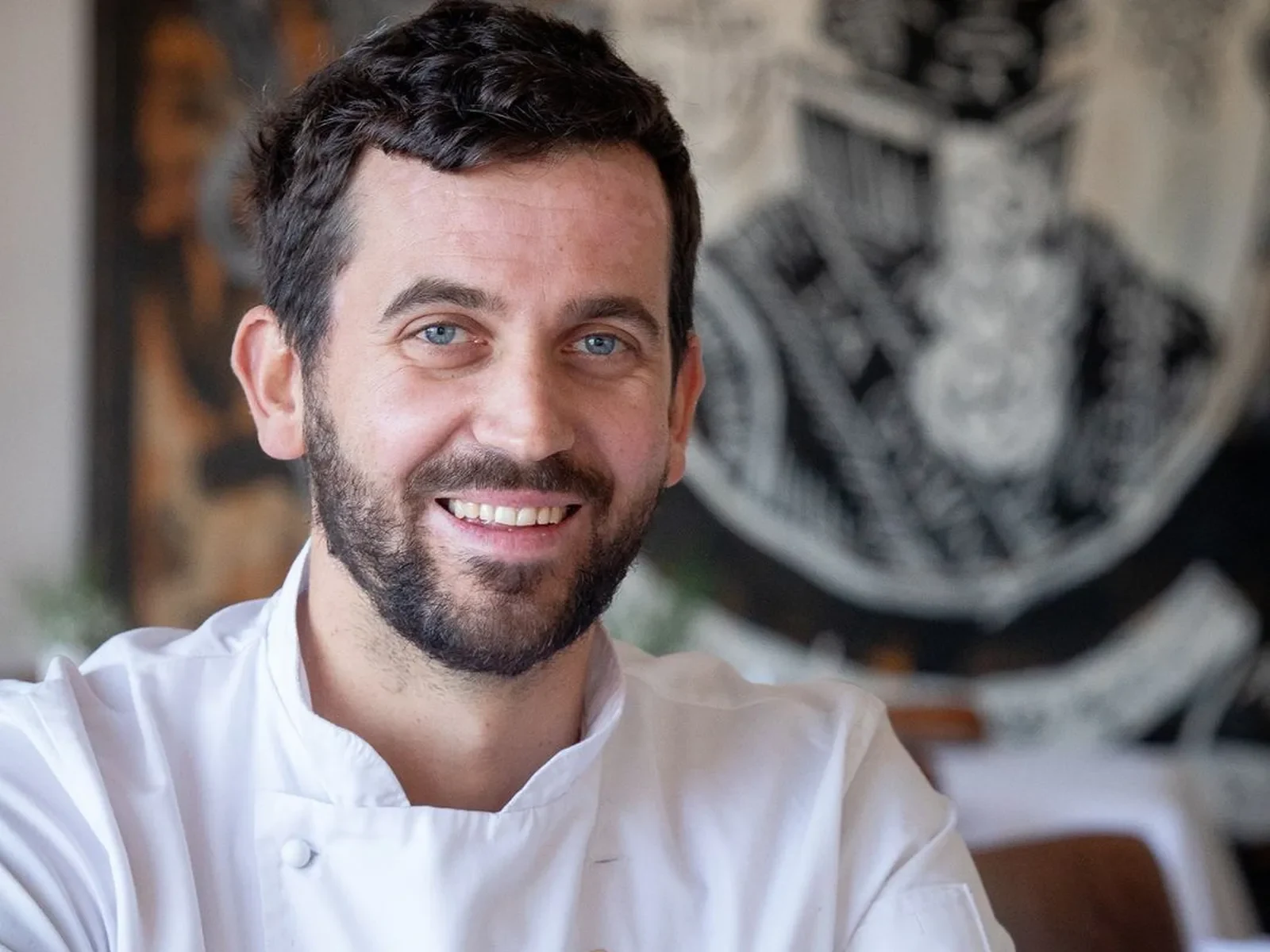 Chef in a white coat smiling in a restaurant setting with artwork behind.