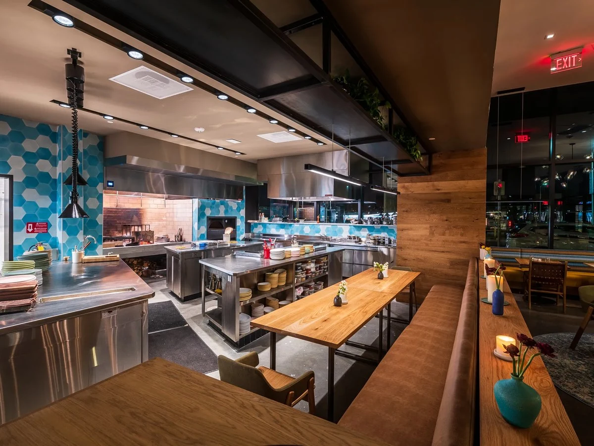 Interior of a contemporary restaurant kitchen and dining area with professional cooking stations, wooden furniture, and blue geometric wall tiles.