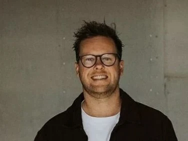 Brendan Pratt, wearing glasses and a dark jacket over a white t-shirt, smiles while standing in an industrial-style room with concrete walls.