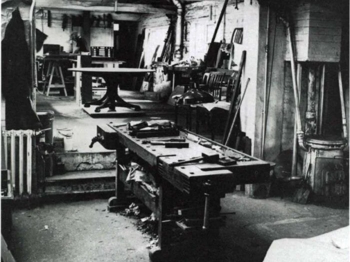 A black-and-white photo of the Edward Barnsley Workshop bench room, showing a workbench with tools and wood shavings, and furniture in the background.