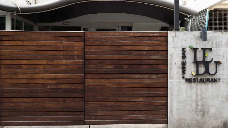 Exterior view of a restaurant entrance with horizontal wooden slat gates and a concrete pillar displaying the Saam SoMA restaurant logo.