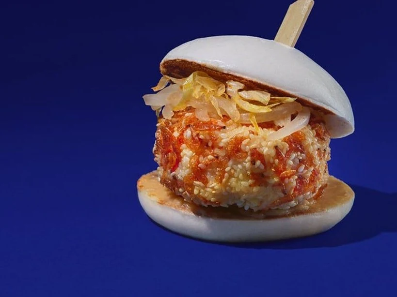 A professional food photograph of a bao (steamed bun) sandwich containing a breaded prawn patty with shredded vegetables, presented on a blue backgrou