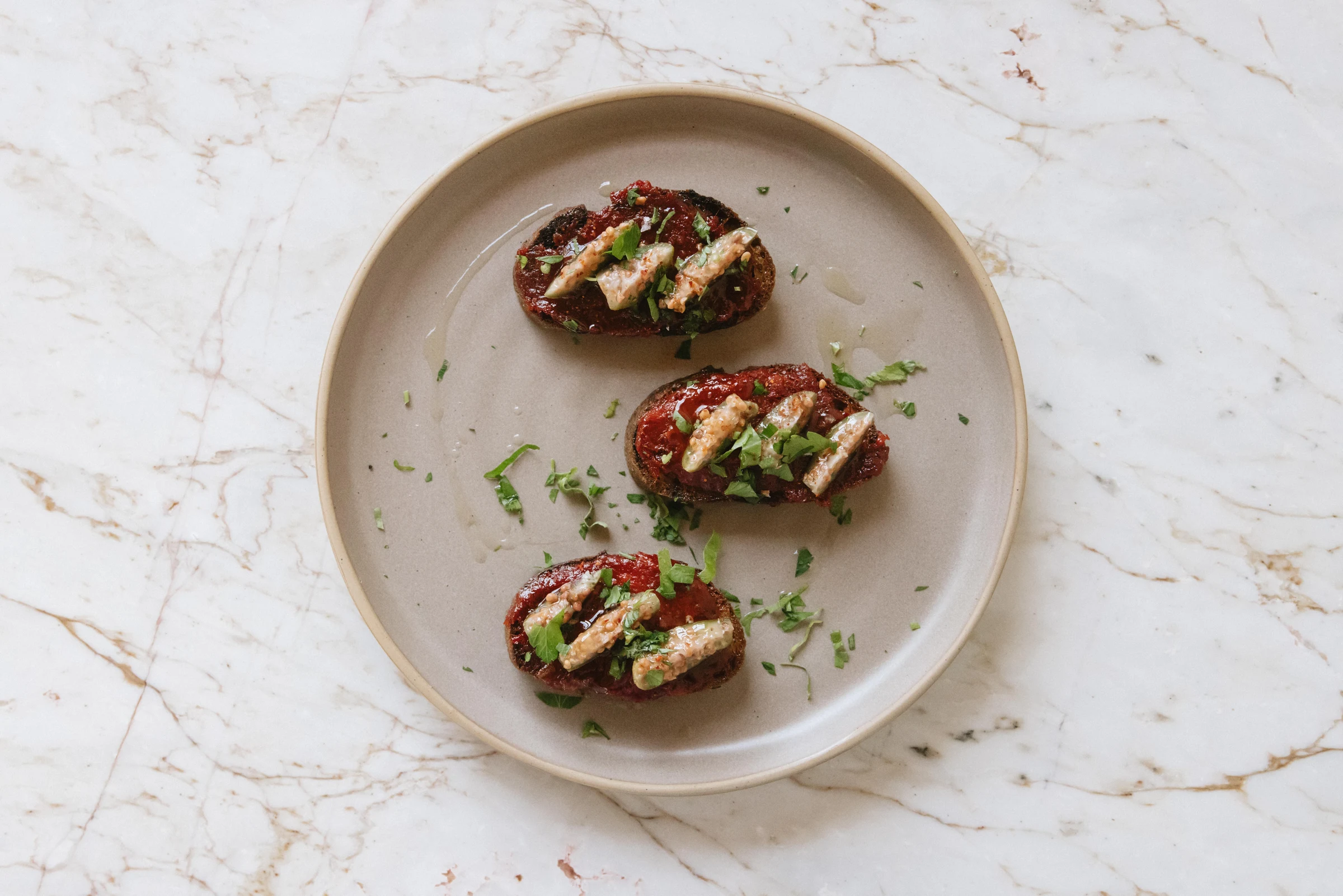 Three crostini appetizers on a beige ceramic plate, garnished with herbs