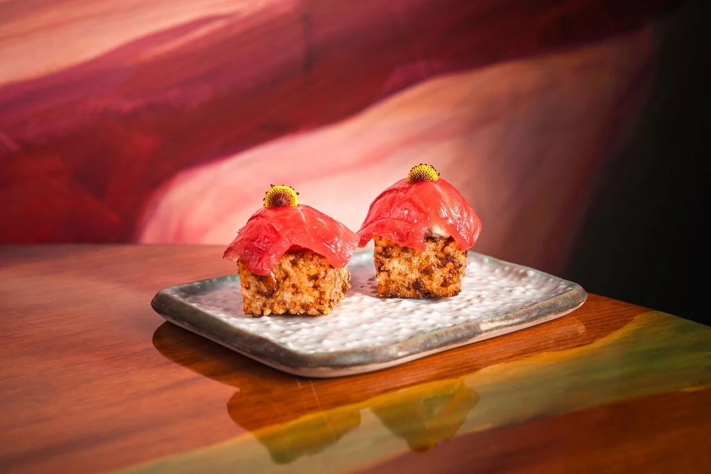 Two small golden cakes with red petal toppings and gold leaf garnish sit on a white plate against a warm-toned blurred background.