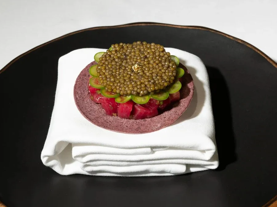 An elegantly plated tasting menu course featuring caviar atop sliced green vegetables and diced red fish, presented on a purple disc on a white napkin.