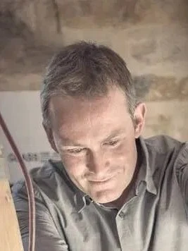 A man with short gray hair wearing a gray shirt concentrates on painting with a brush in his hand.