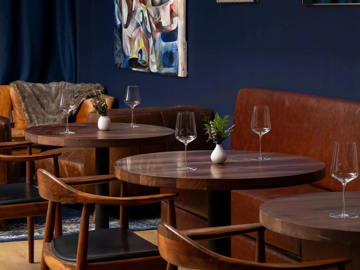 The Saison wine bar interior with round wooden tables, leather banquettes, wine glasses, flowers, and contemporary art.