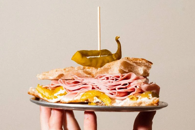 A mortadella sandwich on a metal plate held by a hand, featuring layers of pink mortadella, yellow peppers, and pickled peppers on bread with a toothp