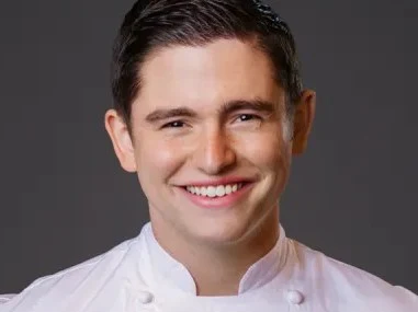 A young man with dark hair, wearing a white chef's coat with green embroidered details, smiles at the camera against a gray background.