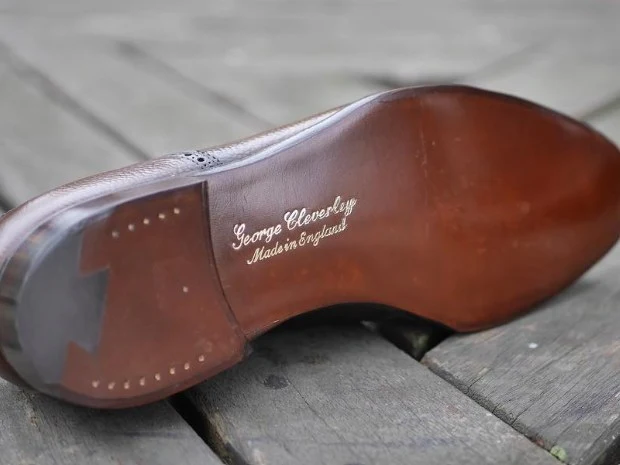 The brown leather sole of a George Cleverley shoe, with 'George Cleverley / Made in England' stamped in gold, rests on wooden planks.
