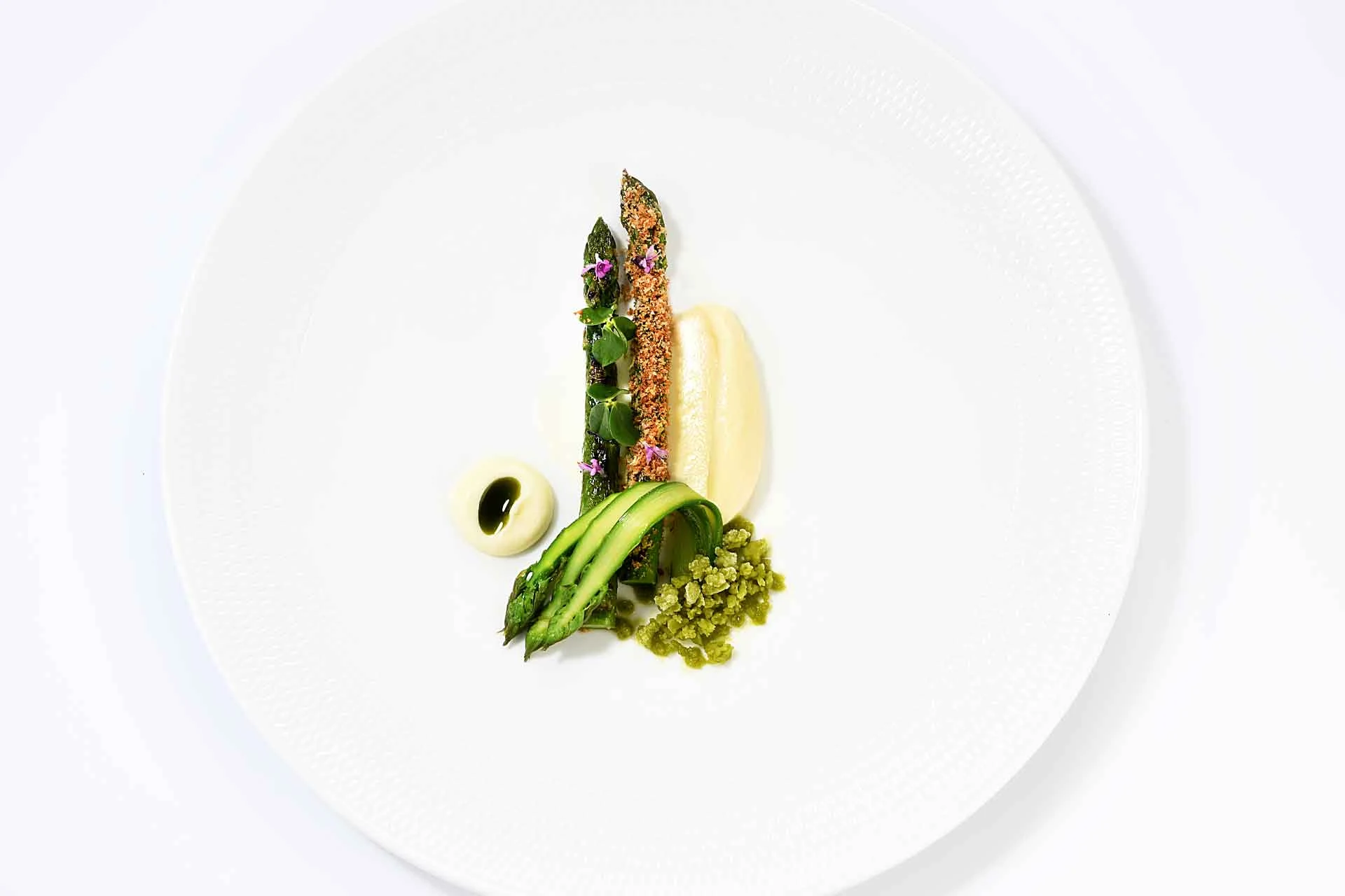 An artfully plated fine-dining dish featuring asparagus with a crusted coating, white puree, avocado slices, and edible flowers on a white plate; the 