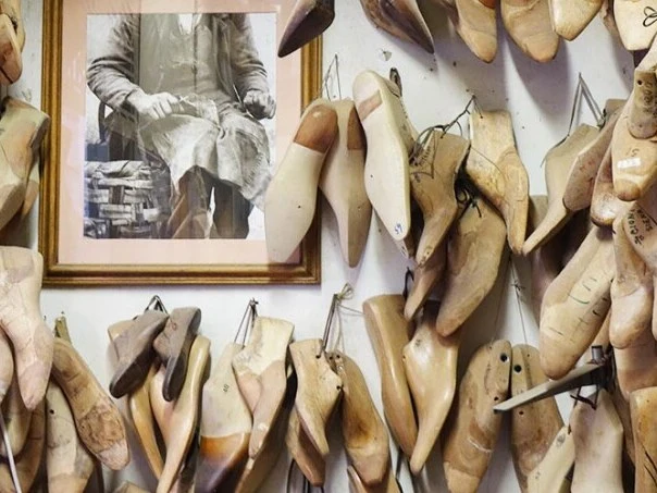 A wall in a shoe workshop is covered with dozens of wooden shoe lasts of various sizes and shapes, with a framed black-and-white photo of a cobbler
