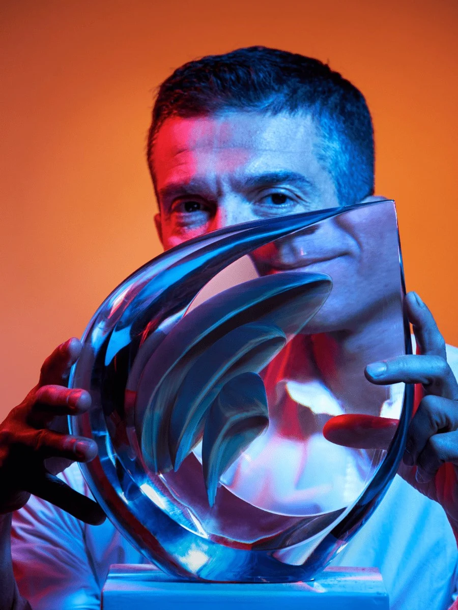 A man, Simone Cenedese, holds a sculptural glass piece in front of his face, illuminated by dramatic blue and red lighting against an orange