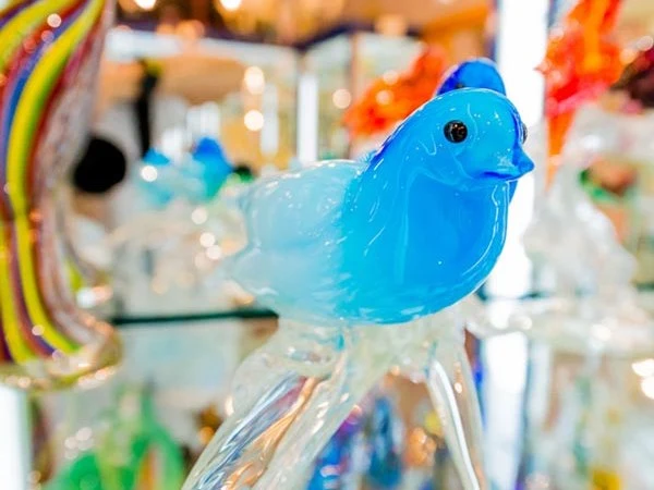 A close-up of a blue Murano glass bird figurine with a clear glass base, displayed in a brightly lit showroom.
