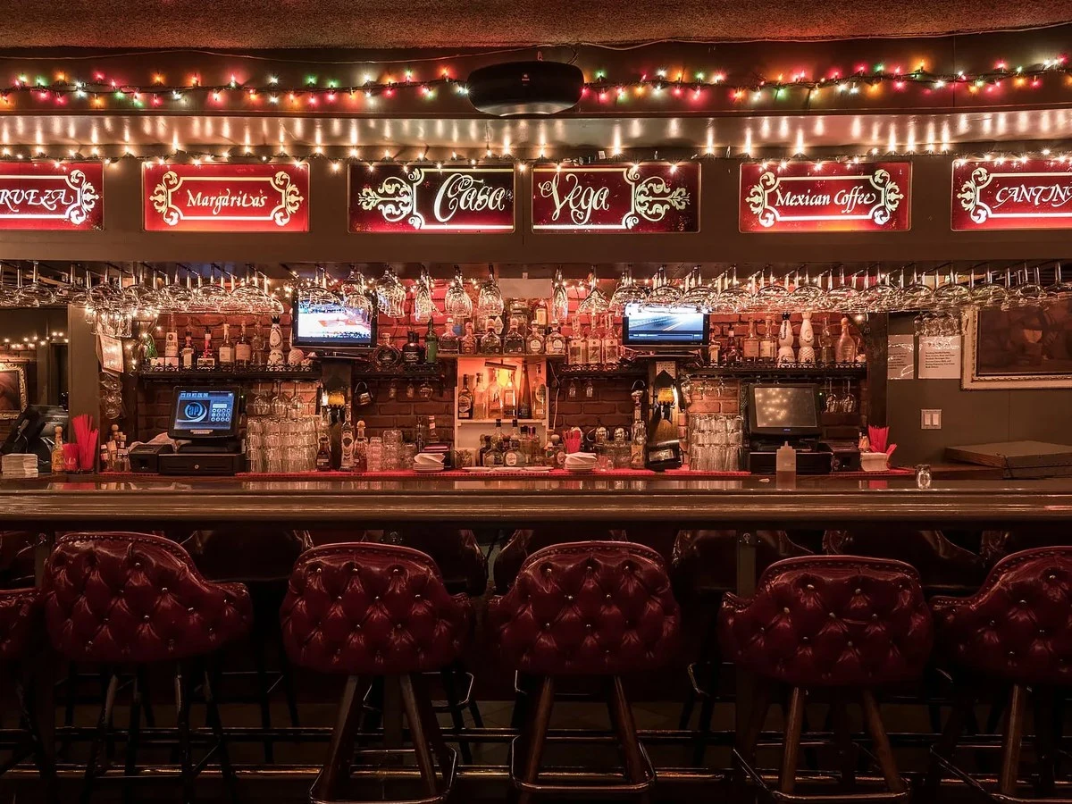 A well-appointed bar interior with red tufted bar stools, extensive back bar with glassware and spirits, decorative signage for beverages (Margaritas,