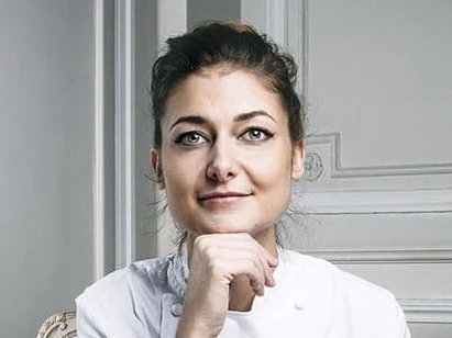 Chef in a white coat, seated in a well-lit room with decorative walls.