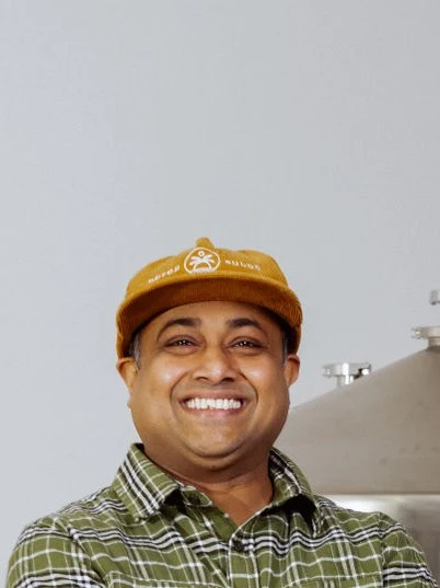A smiling man with arms crossed wearing a green plaid shirt and an orange/mustard cap stands in front of a large stainless steel brewing/distilling ve