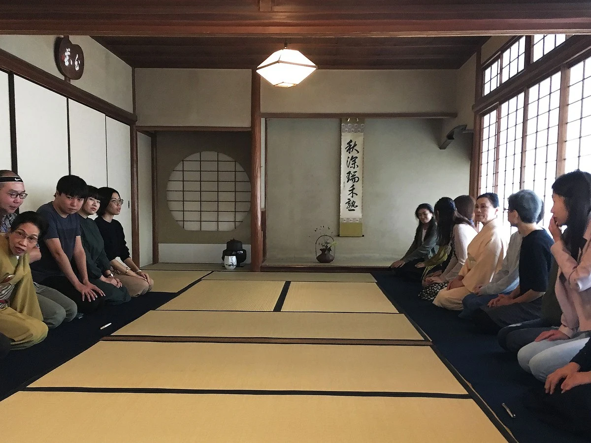 A group of participants sits in seiza on tatami mats in a traditional Japanese tea room, featuring shoji screens and a hanging scroll.