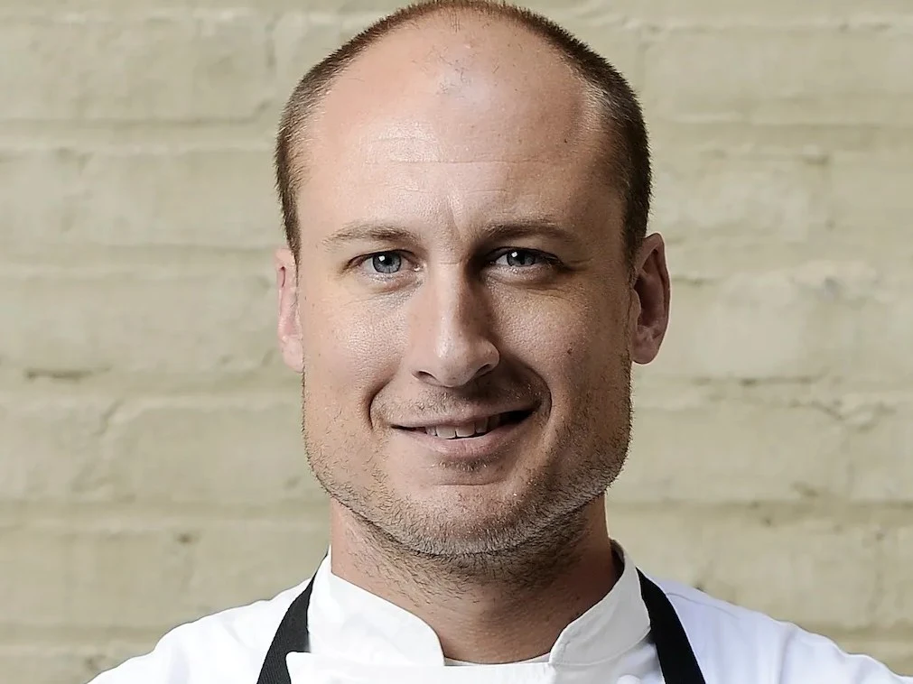 A portrait of chef Dave Beran, a bald man with blue eyes and a slight smile, wearing a white chef's jacket and a black apron against a light-colored.