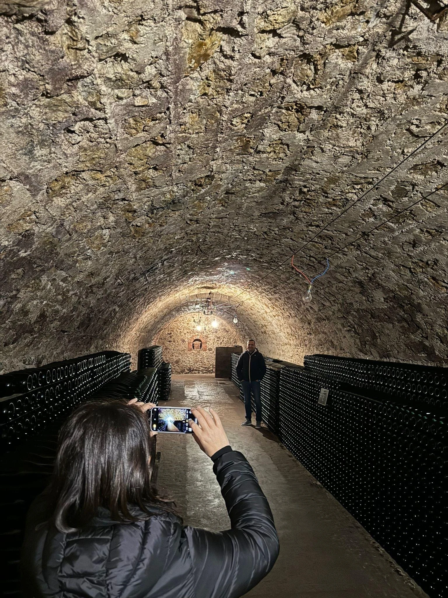 A person photographs another person in a dimly lit, arched Champagne cellar with chalk walls and rows of riddling racks.