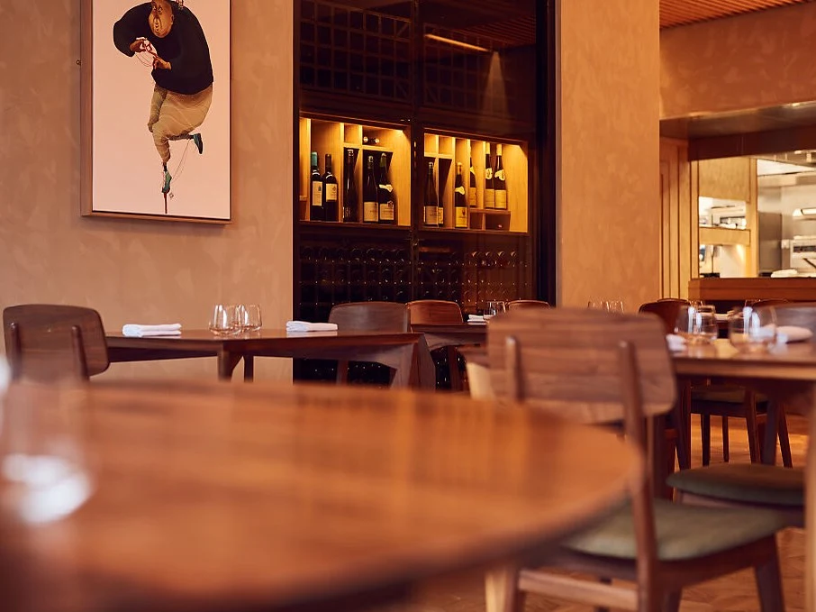 A modern restaurant interior with wooden tables and chairs, a wine storage wall, and an open kitchen where a chef is working.
