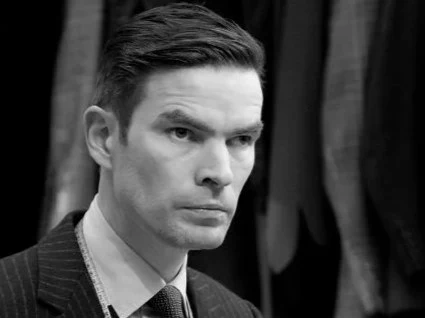 Black and white photograph of a man wearing a dark pinstriped suit jacket and tie, shown in profile against a dark background.
