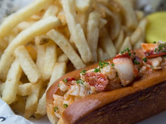 A close-up of a lobster roll with chunks of lobster meat and chives on a toasted bun, served with a side of golden french fries and a yellow lemon.
