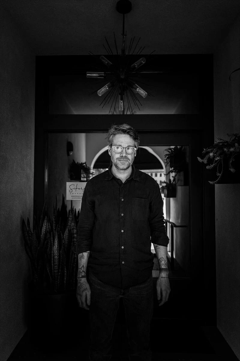 Black and white portrait of Marcus Jernmark, a man with glasses and tattoos, wearing a black button-down shirt, standing in a doorway.