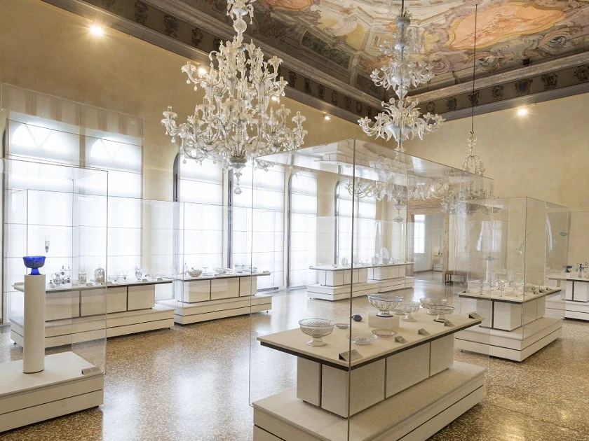 An ornate museum gallery with high ceilings, chandeliers, and multiple glass display cases showcasing various historic glass objects.