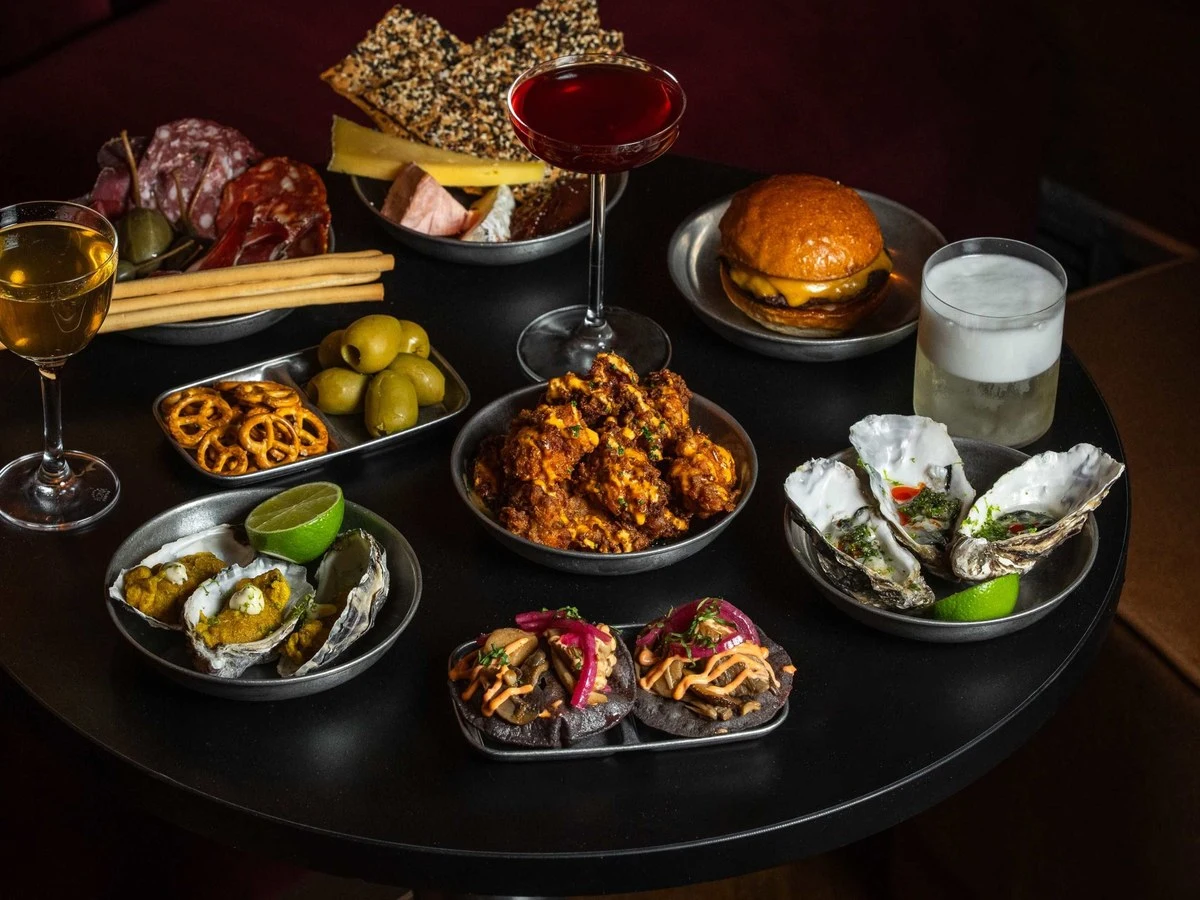 A dark round table laden with various dishes and drinks: charcuterie, fried chicken, oysters, a burger, pretzels, olives, and cocktails.