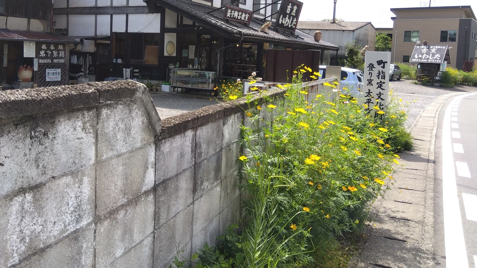 Traditional pottery studios along a street in Mashiko with yellow wildflowers growing beside a concrete wall and shop signs visible.