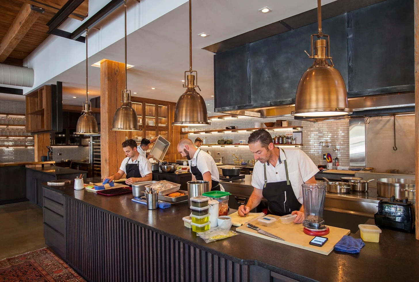 A professional restaurant kitchen with four chefs in white shirts and black aprons working at a large prep island under copper pendant lights; the sou