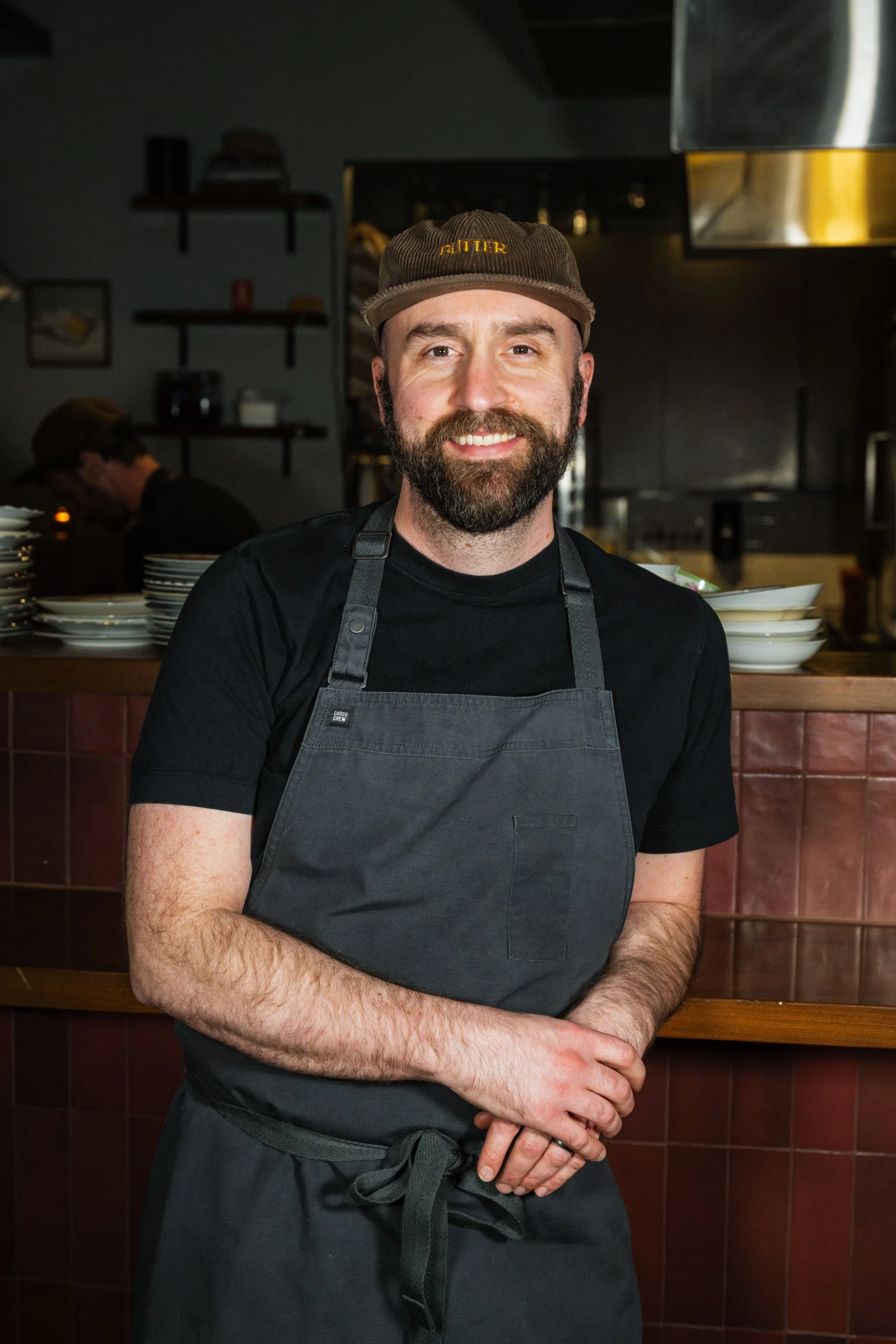 A chef in black apron and corduroy cap stands in a professional kitchen; the source page (StarChefs profile) explicitly attests this is Chef Benjamin 
