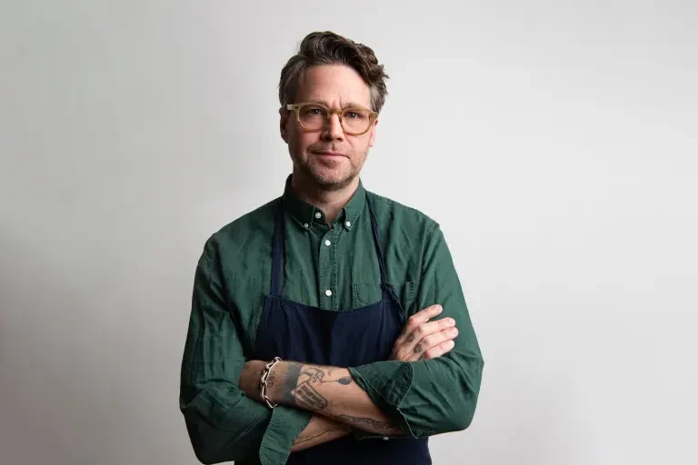 Professional portrait of a chef in green button-down shirt and dark apron with visible tattoos and glasses, shot against a neutral background; Los Ang