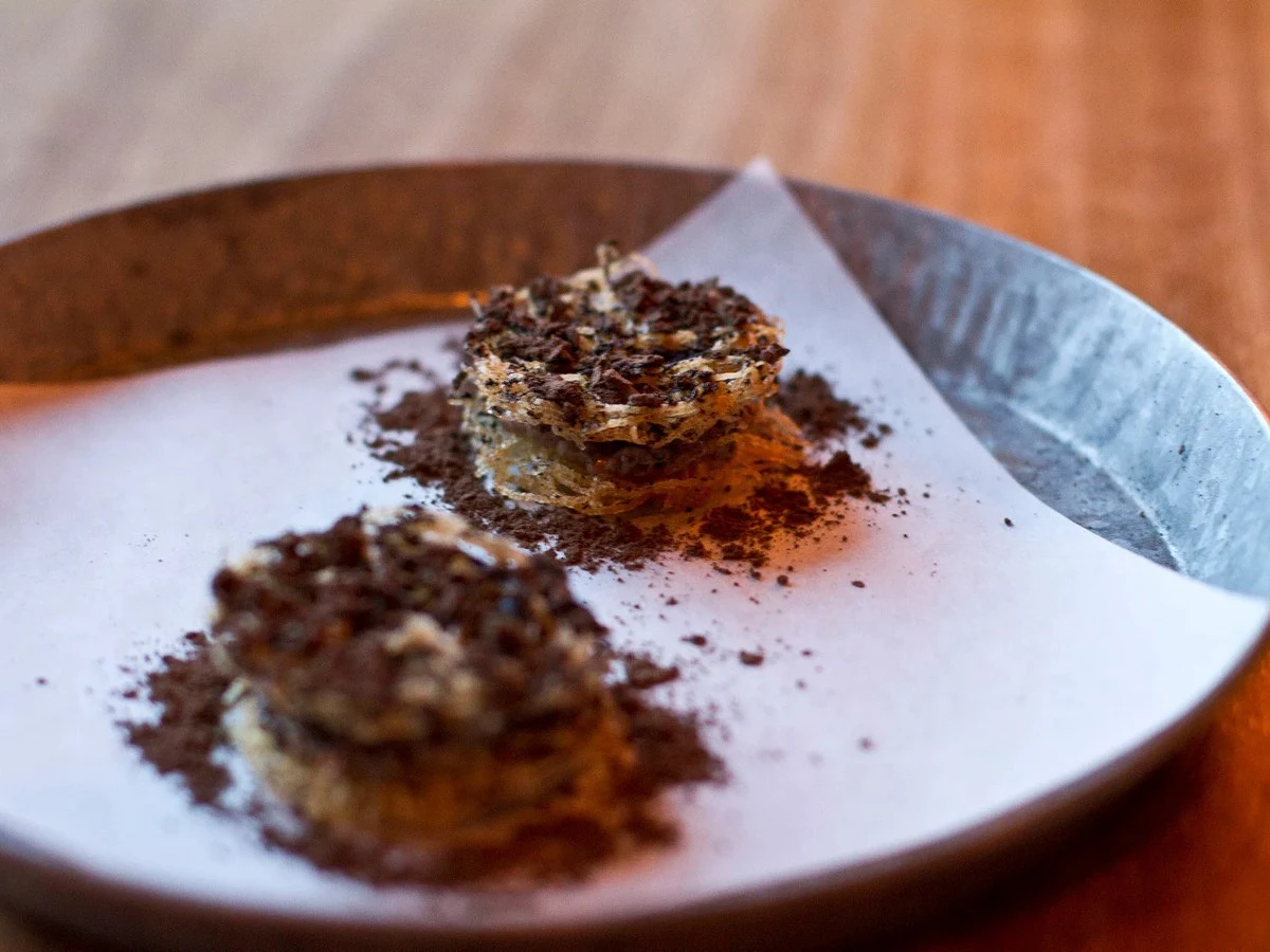 Two small fried potato haystack rounds, dusted with dark powder, are presented on a white paper on a ceramic plate.