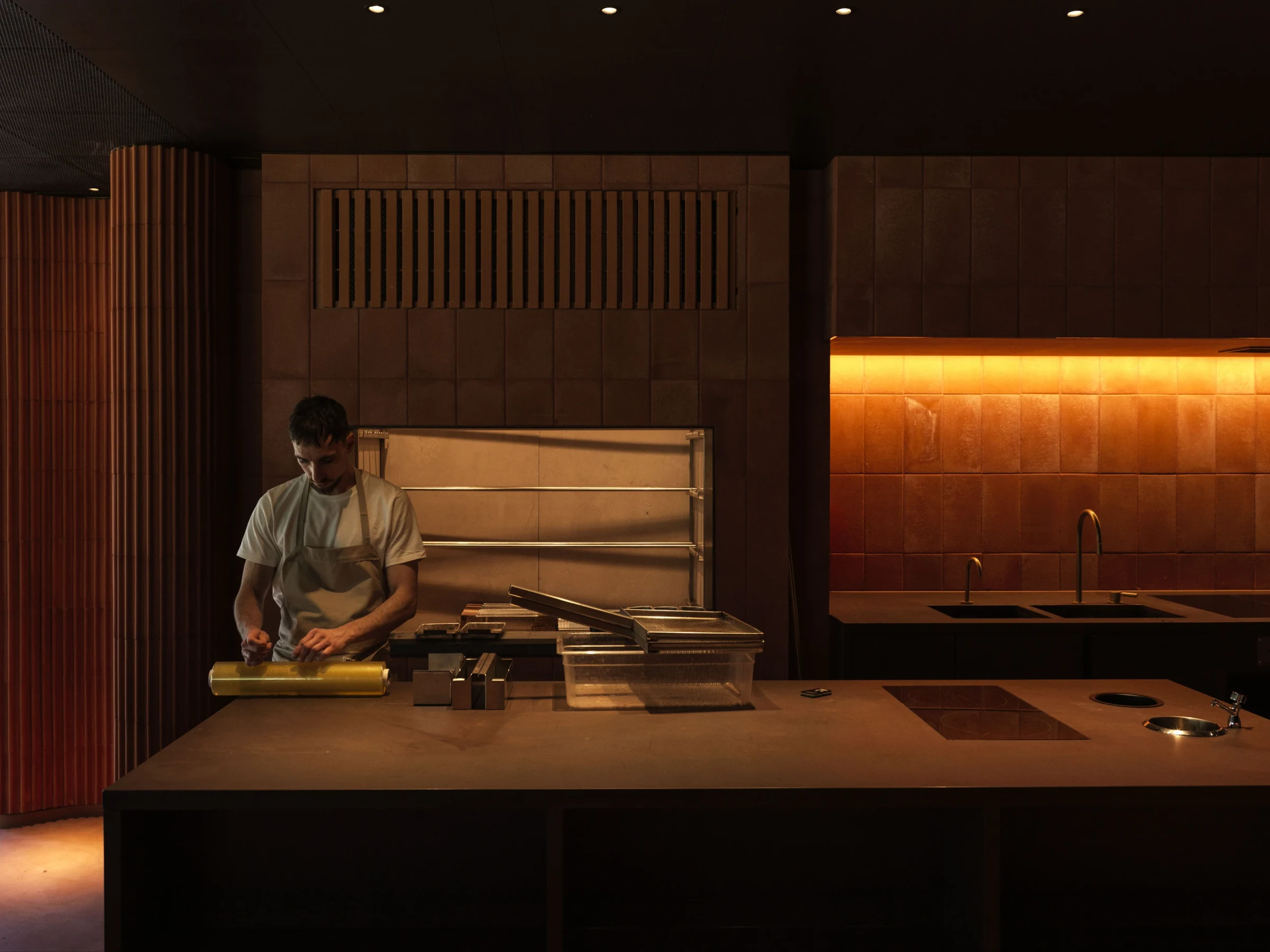 A person wearing an apron and light-colored shirt works at a wooden prep counter in a modern kitchen with warm lighting, wooden wall panels, and organized cooking equipment.
